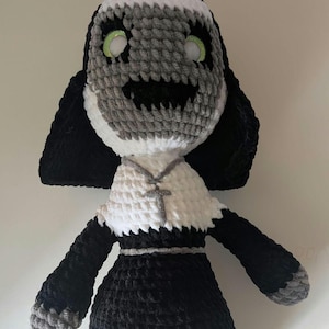 Puede incluir: Un juguete de peluche tejido a crochet con forma de monja con hábito blanco y mangas negras. El juguete tiene una cara gris y blanca con una amplia sonrisa y ojos verdes.