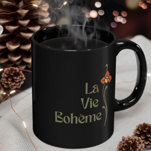May include: A black ceramic mug with the words "La Vie Bohème" in green script, accompanied by a floral design. The mug is filled with a dark liquid, and steam rises from it. The mug is on a light gray surface.