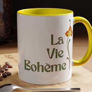 May include: White ceramic mug with a yellow interior and handle. The mug features the text "La Vie Bohème" in green, with a floral design. A spoon and a macaron are on a wooden surface.