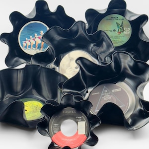 May include: A collection of black, wavy-edged bowls crafted from vinyl records. The bowls display the original record labels, including bands like Queen and Pink Floyd. The labels feature various colors and text, adding visual interest to the unique, upcycled decor.