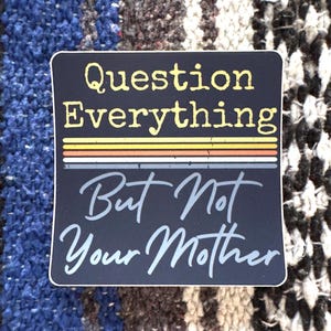 May include: Black and white sticker with the text "Question Everything But Not Your Mother" in a retro style font. The text is in a white box with a black background. There are colorful stripes behind the text.