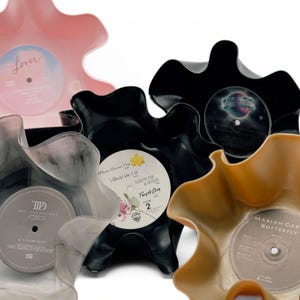 May include: A collection of decorative bowls crafted from repurposed vinyl records. The bowls vary in color, including pink, black, and gold, and feature the original record labels. The edges of the records are shaped into wavy, organic forms.