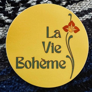 May include: A yellow round sticker with the text "La Vie Bohème" in green and brown lettering. A stylized green flower with a red center is on the right side of the sticker.