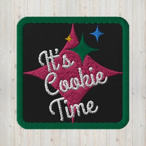 May include: Embroidered patch with the phrase "It's Cookie Time" in white script. The text is centered on a black background, framed by a green border. A pink starburst design is behind the text, with green, yellow, and blue star accents.