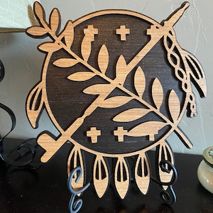 May include: A wooden wall decor featuring a Native American shield design with a cross pattern, a branch with leaves, and a spear. The shield is brown with a black background and is mounted on a black metal stand.