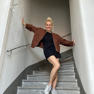 K&ouml;nnte beinhalten: Eine Frau in einer braunen Jacke, einem schwarzen Rock und wei&szlig;en Turnschuhen steht auf einer Betontreppe.