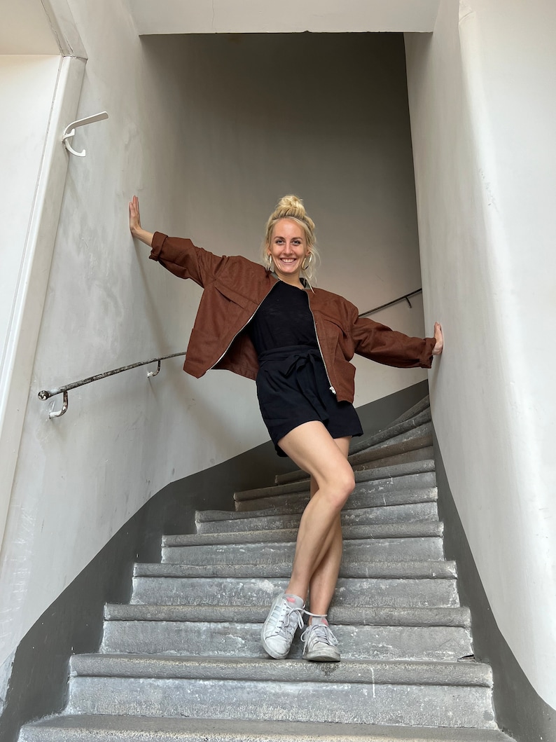 K&ouml;nnte beinhalten: Eine Frau in einer braunen Jacke, einem schwarzen Rock und wei&szlig;en Turnschuhen steht auf einer Betontreppe.