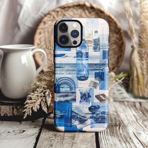 May include: A phone case featuring a blue and white collage design. The design incorporates images of bottles, the sea, and architectural details. The case rests on a wooden surface, with a white jug and dried plants visible in the background.
