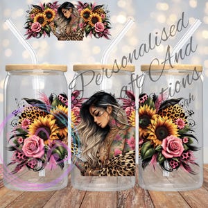 May include: Clear glass can-shaped tumbler with a bamboo lid and a straw. The tumbler features a design of a woman with tattoos, sunflowers, pink roses, and leopard print accents. The background is a bokeh of lights.