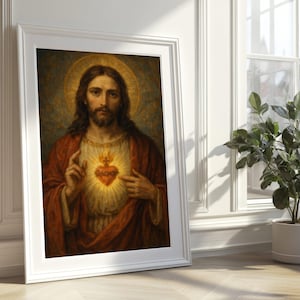 May include: Framed religious artwork depicting a figure with long brown hair, a beard, and a red robe. The figure has a glowing heart on their chest and a golden halo. The artwork is in a white frame and leans against a white wall.