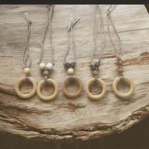 May include: Five necklaces featuring light-colored wooden rings suspended from grey cord. Each necklace includes a unique arrangement of small wooden beads in shades of cream, tan, brown, and dark grey, attached to the ring with a knotted cord loop.