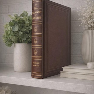 May include: A hardcover book titled "The New Dictionary of Thoughts" in a brown leather-like cover. The spine has gold lettering and decorative elements. The book is displayed on a shelf with other books and decorative items.