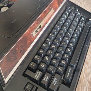 May include: A black Brother Correct-O-Ball typewriter featuring a QWERTY keyboard layout with black keys and white lettering. The device includes a decorative brown wood-grain panel above the keys and a power switch labeled off.