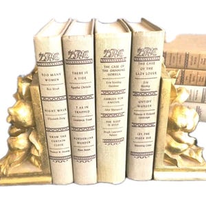 May include: A set of vintage books with off-white covers and gold lettering, held by a gold-colored bookend. The books include titles like "Too Many Women" and "The Case of the Grinning Gorilla."