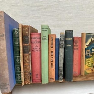 May include: A collection of vintage books in various colors and sizes, including titles like "Marriage of Harlequin" and "Nemesis?" displayed on a wooden book stand. The books have aged covers and spines, creating a classic aesthetic.