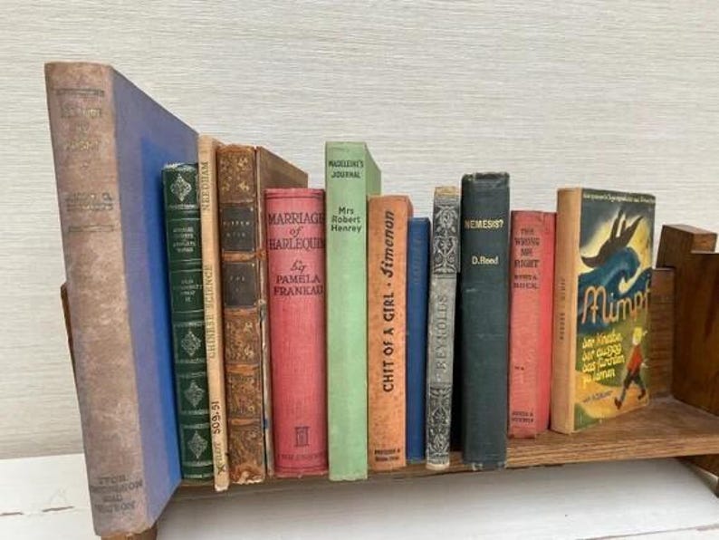May include: A collection of vintage books in various colors and sizes, including titles like "Marriage of Harlequin" and "Nemesis?" displayed on a wooden book stand. The books have aged covers and spines, creating a classic aesthetic.