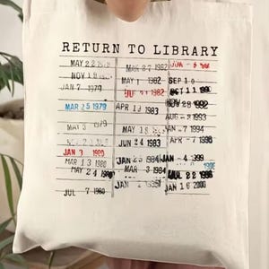 May include: Cream-colored tote bag with the text "RETURN TO LIBRARY" and a vintage library card design printed on it. The design includes dates and stamps, giving it a retro aesthetic. The bag is held up by a person.