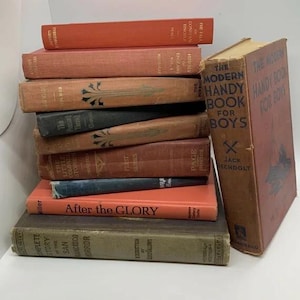 May include: A stack of antique books in various colors, including orange, brown, and dark green. The titles are visible, with one book titled "The Modern Handy Book for Boys." The books are arranged on a white surface.