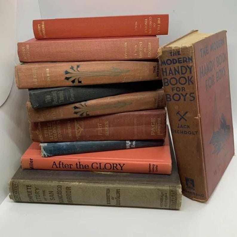 May include: A stack of antique books in various colors, including orange, brown, and dark green. The titles are visible, with one book titled "The Modern Handy Book for Boys." The books are arranged on a white surface.