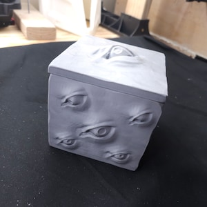 May include: A gray, cube-shaped box with a textured surface and multiple eyes sculpted on the sides. The box has a lid with a single eye sculpted on top.