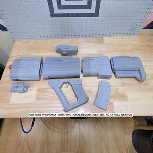 May include: A disassembled gray costume prop gun with various components, including a handle, magazine, and scope. The parts are arranged on a wooden surface. The text "COSTUME PROP ONLY - NON-FUNCTIONAL DECORATIVE ITEM - NOT A REAL WEAPON" is visible.