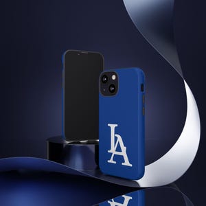 Könnte beinhalten: Eine königsblaue Handyhülle mit den weißen Buchstaben "LA" wird neben einem schwarzen Smartphone gezeigt. Die Hülle hat eine matte Oberfläche und einen erhöhten Rand um die Kamera. Der Hintergrund ist dunkelblau mit einem silbernen Band.