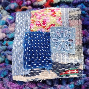 Pode incluir: Close-up de uma obra de arte têxtil com um design de patchwork. A peça apresenta vários quadrados de tecido em tons de azul, rosa e padrões florais, costurados com linha branca. O fundo é um fio texturizado multicolorido.