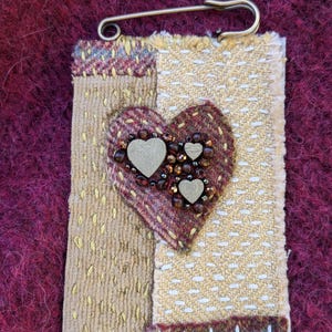 Puede incluir: Un broche de tela hecho a mano con un diseño de corazón. El corazón está hecho de tela marrón y roja y está decorado con cuentas marrones y pequeños corazones dorados. El broche está sujeto a un imperdible.