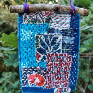 May include: A handcrafted textile wall hanging with a patchwork design. The piece features diverse fabric patterns in blue, red, and green, with a sleeping fox applique. It is suspended from a small wooden branch with colourful twine.