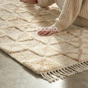 Puede incluir: Una alfombra rectangular beige con un patrón geométrico y bordes con flecos. La alfombra tiene un aspecto texturizado y está hecha de un material de fibra natural. El diseño presenta un patrón repetitivo de cuadrados y diamantes.
