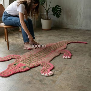 May include: A pink and green alligator-shaped rug with a textured design. The rug features a detailed pattern, including the alligator's scales, eyes, and feet. The rug is placed on a concrete floor.