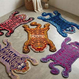 Puede incluir: Cinco alfombras con forma de tigre en tonos rosa, naranja, azul, morado y lavanda. Cada alfombra presenta un diseño detallado de la cara y el cuerpo del tigre. Las alfombras están colocadas sobre un suelo gris, con una cesta tejida y objetos decorativos en el fondo.