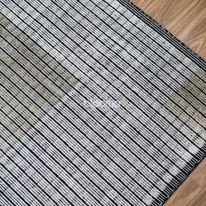 May include: A close-up of a rug with a geometric pattern. The rug features alternating stripes of black and off-white, creating a textured appearance. The rug is placed on a wooden floor.