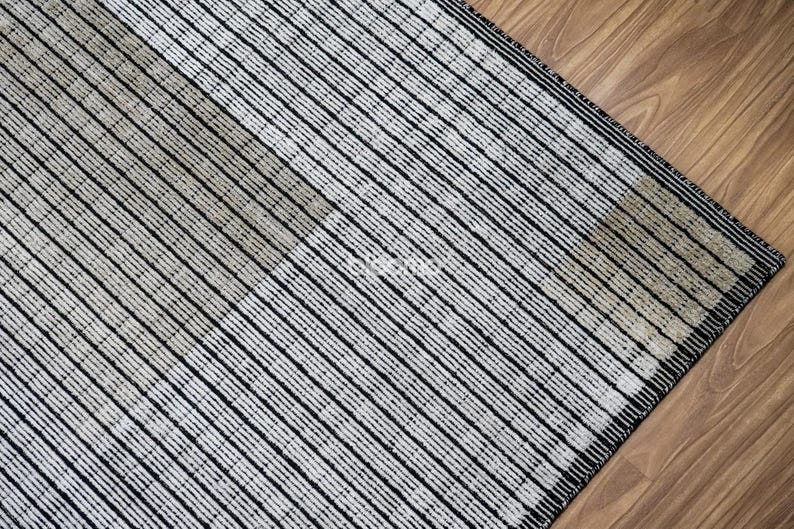 May include: A close-up of a rug with a geometric pattern. The rug features alternating stripes of black and off-white, creating a textured appearance. The rug is placed on a wooden floor.