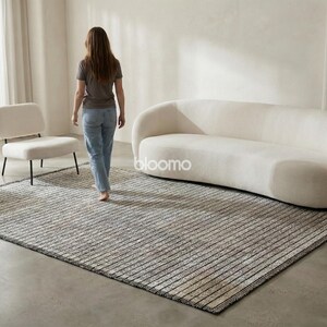 May include: A rectangular area rug with a striped pattern in shades of gray and beige. The rug is placed on a concrete floor, with a white sofa and two chairs in the background. The word "bloomo" is visible on the sofa.