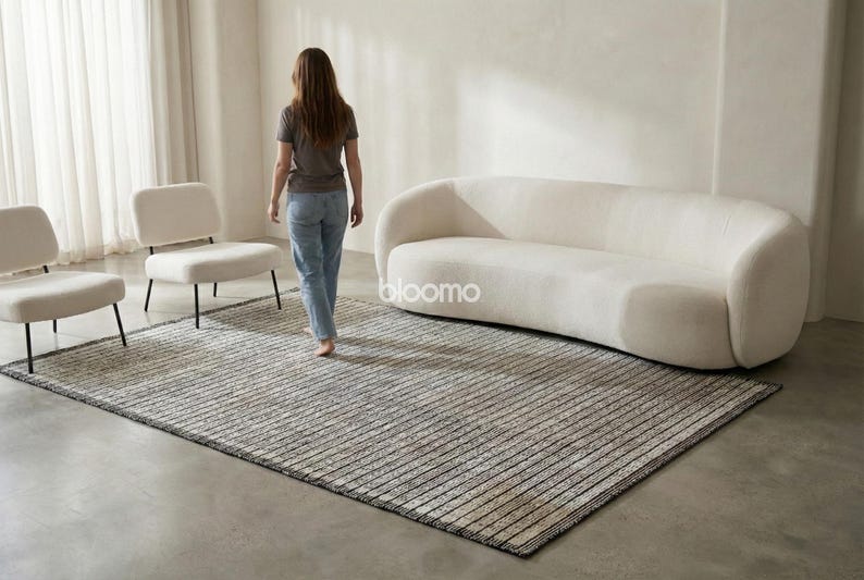 May include: A rectangular area rug with a striped pattern in shades of gray and beige. The rug is placed on a concrete floor, with a white sofa and two chairs in the background. The word "bloomo" is visible on the sofa.