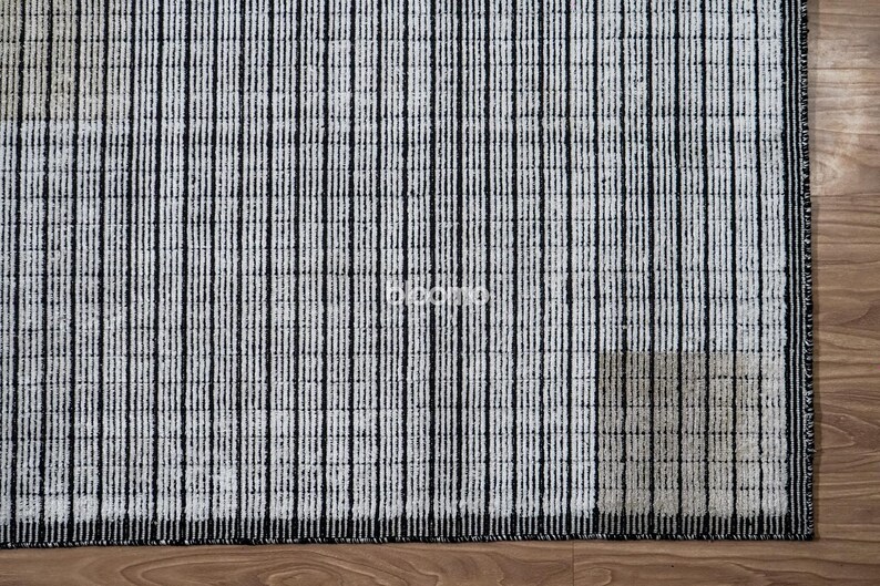 May include: A rectangular rug with a pattern of vertical black stripes on a white background. The rug is made of a textured material and is placed on a wooden floor. The rug's design is simple and modern.