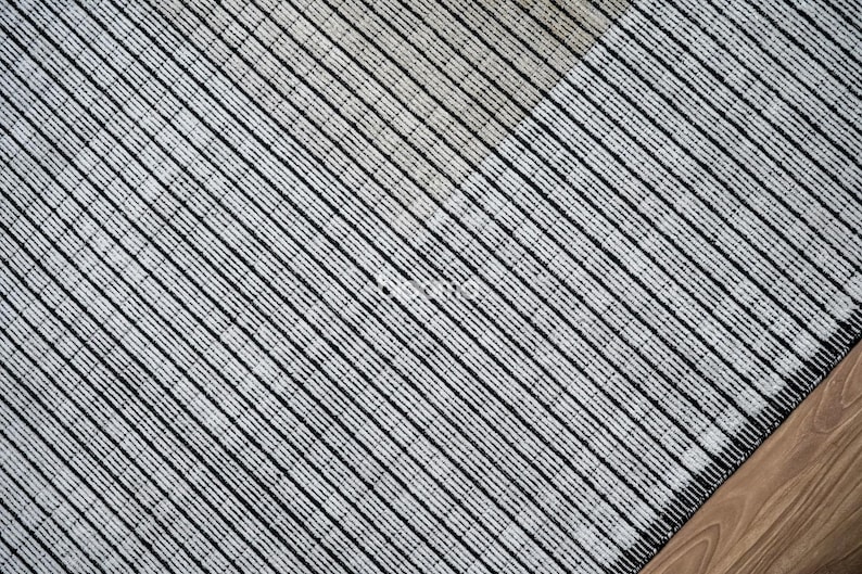 May include: Close-up of a textured rug with a diagonal striped pattern in black and white. The rug's corner is visible, resting on a wooden floor.