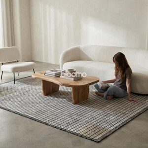 May include: A modern living room setup featuring a cream-colored sofa, two armchairs, and a wooden coffee table. A striped rug covers the floor. A person is sitting on the rug, reading a book. The room has a minimalist design.