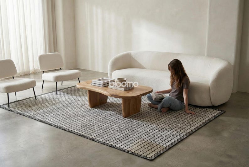 May include: A modern living room setup featuring a cream-colored sofa, two armchairs, and a wooden coffee table. A striped rug covers the floor. A person is sitting on the rug, reading a book. The room has a minimalist design.