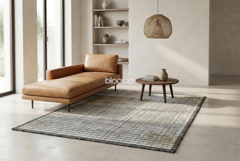 May include: A modern living room scene featuring a tan leather chaise lounge, a round wooden coffee table, and a patterned rug. A woven pendant light hangs above the table. Neutral tones and natural light create a serene atmosphere. Decorative vases and books are on display.