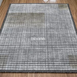 May include: Rectangular area rug with a striped pattern in shades of gray and black. The rug has a fringe border and is placed on a wooden floor. The word "bloom" is visible on the rug.