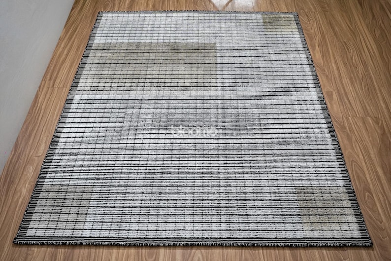 May include: Rectangular area rug with a striped pattern in shades of gray and black. The rug has a fringe border and is placed on a wooden floor. The word "bloom" is visible on the rug.