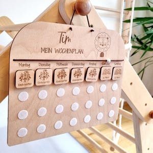 Könnte beinhalten: Ein wöchentliches Holz-Planungstableau mit dem Namen "Tim" oben. Das Tableau hat die Wochentage in deutscher Sprache beschriftet, mit Platz für Sticker oder Magnete. Das Tableau zeigt eine Löwenillustration und den Text "Mein Wochenplan".