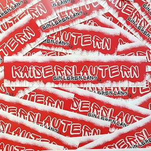 Könnte beinhalten: Ein Stapel roter und weißer Aufkleber mit dem Text "Kaiserslautern Ein Leben Lang" in schwarzer Schrift. Die Aufkleber sind mit einem weißen Spritz-Effekt bedruckt.