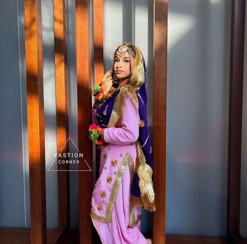 Op de afbeelding: Een vrouw in een roze en paarse outfit met gouden borduurwerk en een paarse dupatta. Ze staat voor een houten muur.