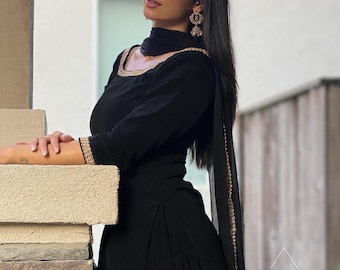 May include: A black salwar kameez ensemble with gold trim on the neckline, sleeves, and dupatta. The outfit includes a long-sleeved tunic and matching pants. The model is wearing gold earrings.