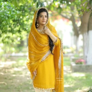 Puede incluir: Una mujer vistiendo un salwar kameez amarillo con bordados dorados y un dupatta amarillo a juego. Est&aacute; de pie en un entorno de jard&iacute;n.