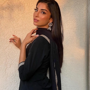 Könnte beinhalten: Eine Frau in einem schwarzen Salwar Kameez, einem traditionellen südasiatischen Outfit. Das Outfit hat goldene Stickereien an den Ärmeln und Kanten des Dupatta. Sie trägt auch silberne Ohrringe.