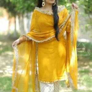 Puede incluir: Una mujer lleva un salwar kameez amarillo con un dupatta blanco y pantalones blancos. El atuendo tiene bordados dorados y un ribete con volantes. La mujer est&aacute; de pie en un campo de hierba.